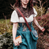 Chloé est une jeune femme à la peau claire. Elle avance d'un pas décidé et enjoué vers le côté droit de l'image. Ses cheveux aubruns lui arrivent juste au dessus des épaules. Dans sa main droite, elle tient un livre ancien. Elle porte une blouse blanche à manches courtes sur laquelle se trouve un boléro en simili-cuir brun, fermé par de jolis boutons en laiton vieil or. Elle porte également une jupe dont la partie corsetée commence sous sa poitrine et arrive jusqu'au dessus de ses hanches. Cette partie corsetée est composée d'une alternance de panneaux en lin bleu canard et de coton bio à motifs de fleurs et de champignons, dans un style très doux et poétique. Le reste de la jupe est en lin bleu canard et arrive juste au dessus de ses genoux. Elle porte en bandoulière un sac arrondi assorti à la jupe. Son rabat est réalisé avec le coton à motifs, et le corps du sac est en lin bleu canard. La hanse est en coton marron. Le fond de l'image est composé de buissons et d'arbustes en tout genres. La photo a été prise par la talentueuse Namidaël Photographies.