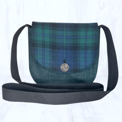 Image carrée sur fond clair. Elle est centrée sur un petit sac arrondi en lin vert foncé, dont le rabat est en tartan vert, bleu foncé et noir. Le rabat du sac est fermé par un bouton rond argenté. Les hanses du sac sont noires.