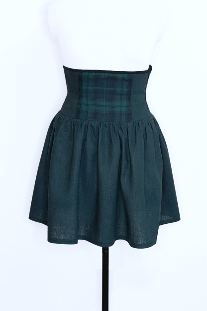 Vue de face de la Jupe Urania Tartan Vert. C'est une jupe dont la partie corsetée commence sous la poitrine et descend jusqu'au dessus du bassin. La partie basse de la jupe est froncée pour donner de l'amplitude et de jolis plis. Elle est faite en lin vert avec certains panneaux de la partie corsetée en tartan vert et noir.