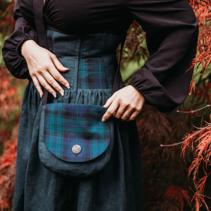 Photographie au format carré d'une jeune femme de profil présentant un sac en lin vert foncé avec un rabat en tartan vert, bleu et noir. La photographie est centrée sur le sac, mais on peut voir qu'elle porte une jupe corsetée réalisée dans avec les mêmes tissus que le sac. On aperçoit sur les côtés de l'images des branches de feuilles d'érables dans les tons rouges.