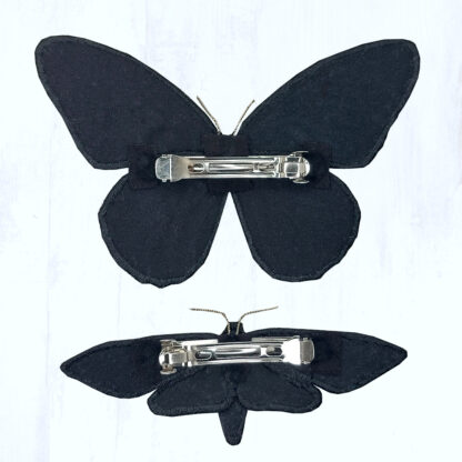 Sur un fond gris clair se trouvent 2 épingles à cheveux en forme de papillon, posées sur le dos afin que l'on puisse voir comment elles s'attachent dans les cheveux. L'envers des papillons est en coton noir, et une grosse barrette en métal argenté est fixée dans le sens de la largeur.