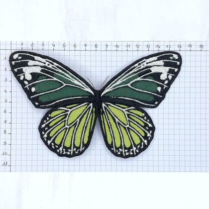 Sur une feuille quadrillée sont notées les graduations en cm dans le sens de longueur et de la hauteur, en partant du coin en haut à gauche. Dessus est posé une barrette à cheveux en forme de papillon vert réalisé en coton brodé. Ce papillon fait environ 16.5 cm de large pour 10.5 cm de hauteur.