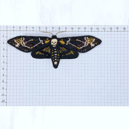 Sur une feuille quadrillée sont notées les graduations en cm dans le sens de longueur et de la hauteur, en partant du coin en haut à gauche. Dessus est posé une barrette à cheveux en forme de papillon sphinx tête de mort, réalisé en coton brodé. Ce papillon fait environ 14.5 cm de large pour 6 cm de hauteur.