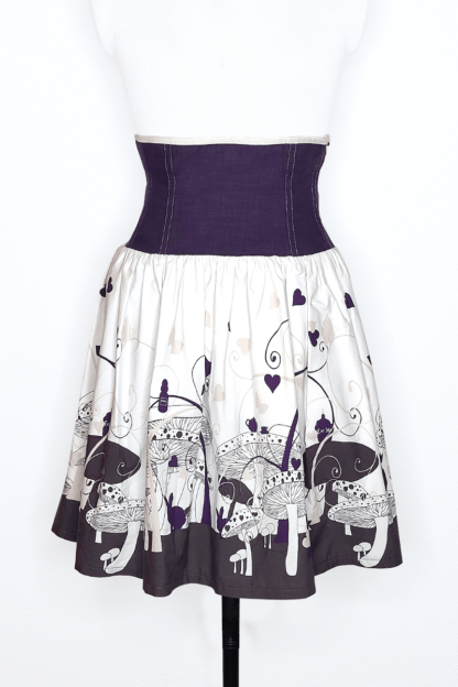 photographie représentant une jupe corsetée vue de face, portée par un mannequin de couture devant un fond blanc. La jupe est composée en 2 parties. Entre le dessous de poitrine et les hanches se trouve une partie corsetée composée de panneaux en lin violet. Cette partie corsetée permet de marquer la taille, tout en étant très confortable. Le bas de la jupe est en coton bio blanc et violet, avec des motifs de champignons, de lapin et de tasse de thé, inspiré de l'univers d'Alice au Pays des merveilles.