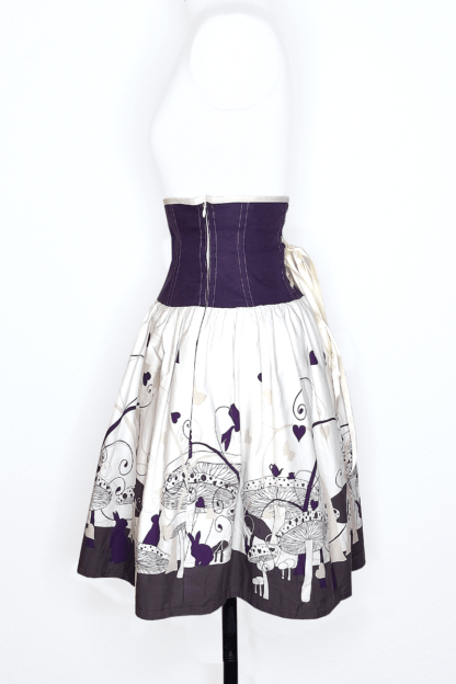 photographie représentant une jupe corsetée vue profil, portée par un mannequin de couture devant un fond blanc. La jupe est composée en 2 parties. Entre le dessous de poitrine et les hanches se trouve une partie corsetée composée de panneaux en lin violet. La jupe s'enfile grâce à une fermeture éclair invisible sur le côté. Cette partie corsetée permet de marquer la taille, tout en étant très confortable. Le bas de la jupe est en coton bio blanc et violet, avec des motifs de champignons, de lapin et de tasse de thé, inspiré de l'univers d'Alice au Pays des merveilles. Invisibles sur la photo, la jupe possède des poches cachées dans les coutures de côtés !