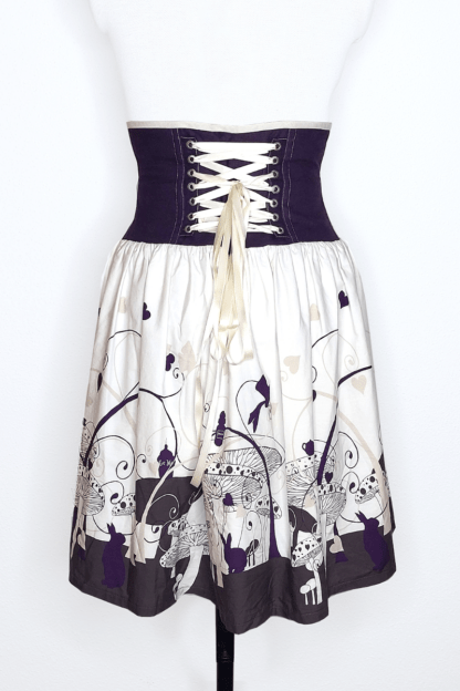 photographie représentant une jupe corsetée vue de dos, portée par un mannequin de couture devant un fond blanc. Entre le dessous de poitrine et les hanches se trouve une partie corsetée composée de panneaux en lin violet. Le milieu du dos est orné d'un laçage en satin blanc cassé, qui, en plus d'être très joli, permet surtout de pouvoir ajuster la jupe à sa morphologie ! Cette partie corsetée permet de marquer la taille, tout en étant très confortable. Le bas de la jupe est coton blanc cassé, dont le bas est chargé de motifs violets inspirés de l'univers d'Alice au Pays des Merveilles. Cette partie est froncée afin d'apporter du volume et de la fluidité.