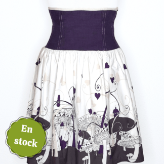 photographie représentant une jupe corsetée vue de face, portée par un mannequin de couture devant un fond blanc. La jupe est composée en 2 parties. Entre le dessous de poitrine et les hanches se trouve une partie corsetée composée de panneaux en lin violet. Cette partie corsetée permet de marquer la taille, tout en étant très confortable. Le bas de la jupe est en coton bio blanc et violet, avec des motifs de champignons, de lapin et de tasse de thé, inspiré de l'univers d'Alice au Pays des merveilles. Sur le côté gauche de l'image se trouve une pastille verte avec écrit "En stock".