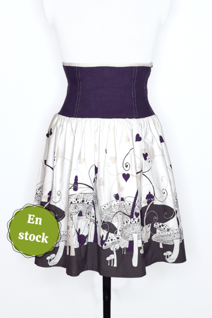 photographie représentant une jupe corsetée vue de face, portée par un mannequin de couture devant un fond blanc. La jupe est composée en 2 parties. Entre le dessous de poitrine et les hanches se trouve une partie corsetée composée de panneaux en lin violet. Cette partie corsetée permet de marquer la taille, tout en étant très confortable. Le bas de la jupe est en coton bio blanc et violet, avec des motifs de champignons, de lapin et de tasse de thé, inspiré de l'univers d'Alice au Pays des merveilles. Sur le côté gauche de l'image se trouve une pastille verte avec écrit "En stock".