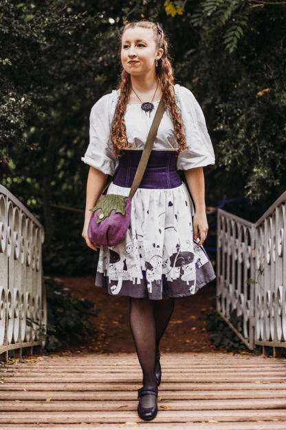 Photographie de la jupe Urania Alice portée. La vue est prise de face. On y voit une jeune femme caucasienne aux longs cheveux blonds marcher sur un pont en bois. La jupe qu'elle porte est composée d'une partie corsetée en lin violet et surmonté d'un biais blanc cassé. Le laçage au milieu dos est également blanc cassé, et permet d'ajuster la jupe à sa morphologie. A partir du bassin, la jupe est en coton blanc à motifs violets inspiré de l'univers d'Alice au Pays des Merveilles. On y voit des champignons, des lapins, des coeurs, des tasses de thé... Les motifs sont principalement placés sur le bas de la jupe, et sont de plus en plus espacés en allant vers le haut de la jupe. Elle porte également un sac Feuille que j'ai réalisé, avec un corps vieux rose et un rabat vert olive.