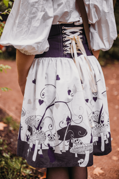 Photographie de la jupe Urania Alice portée. La vue est prise de dos, avec un plan rapproché permettant de voir la jupe dans son intégralité. Elle est composée d'une partie corsetée en lin violet et surmonté d'un biais blanc cassé. Le laçage au milieu dos est également blanc cassé, et permet d'ajuster la jupe à sa morphologie. A partir du bassin, la jupe est en coton blanc à motifs violets inspiré de l'univers d'Alice au Pays des Merveilles. On y voit des champignons, des lapins, des coeurs, des tasses de thé... Les motifs sont principalement placés sur le bas de la jupe, et sont de plus en plus espacés en allant vers le haut de la jupe.
