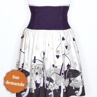 photographie représentant une jupe corsetée vue de face, portée par un mannequin de couture devant un fond blanc. La jupe est composée en 2 parties. Entre le dessous de poitrine et les hanches se trouve une partie corsetée composée de panneaux en lin violet. Cette partie corsetée permet de marquer la taille, tout en étant très confortable. Le bas de la jupe est en coton bio blanc et violet, avec des motifs de champignons, de lapin et de tasse de thé, inspiré de l'univers d'Alice au Pays des merveilles. Sur le côté gauche de l'image se trouve une pastille orange avec écrit "sur commande".