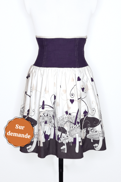 photographie représentant une jupe corsetée vue de face, portée par un mannequin de couture devant un fond blanc. La jupe est composée en 2 parties. Entre le dessous de poitrine et les hanches se trouve une partie corsetée composée de panneaux en lin violet. Cette partie corsetée permet de marquer la taille, tout en étant très confortable. Le bas de la jupe est en coton bio blanc et violet, avec des motifs de champignons, de lapin et de tasse de thé, inspiré de l'univers d'Alice au Pays des merveilles. Sur le côté gauche de l'image se trouve une pastille orange avec écrit "sur commande".