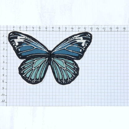 Sur une feuille quadrillée sont notées les graduations en cm dans le sens de longueur et de la hauteur, en partant du coin en haut à gauche. Dessus est posé une barrette à cheveux en forme de papillon vert réalisé en coton brodé. Ce papillon fait environ 13 cm de large pour 8.5 cm de hauteur.