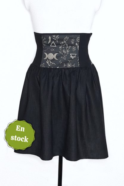 photographie représentant une jupe corsetée vue de face, portée par un mannequin de couture devant un fond blanc. La jupe est composée en 2 parties. Entre le dessous de poitrine et les hanches se trouve une partie corsetée composée d'une alternance de panneaux en lin noir et coton bio noir à motifs ésotériques beige. On peut y voir des sigils, des plantes, des runes, le symbole de la triple lune,... Cette partie corsetée permet de marquer la taille, tout en étant très confortable. Le bas de la jupe est en lin noir. Sur le côté gauche de l'image se trouve une pastille verte avec écrit "en stock".