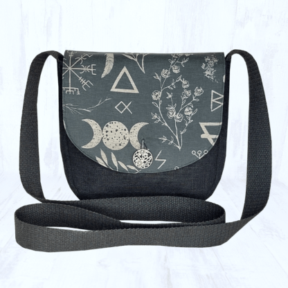 Image carrée sur fond clair. Elle est centrée sur un petit sac arrondi en lin noir, dont le rabat est en coton bio noir à motifs ésotériques beige. On peut y voir des sigils, des plantes, des runes, le symbole de la triple lune,... Le rabat du sac est fermé par un bouton rond argenté. Les hanses du sac sont noires.