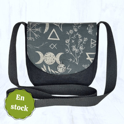 Image carrée sur fond clair. Elle est centrée sur un petit sac arrondi en lin noir, dont le rabat est en coton bio noir à motifs ésotériques beige. On peut y voir des sigils, des plantes, des runes, le symbole de la triple lune,... Le rabat du sac est fermé par un bouton rond argenté. Les hanses du sac sont noires. Dans le coin en bas à gauche, il y a une vignette avec noté "en stock".