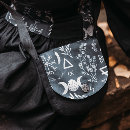 Photographie centrée sur un sac arrondi porté en bandoulière. Son corps est en lin noir, et son rabat est réalisé en coton bio noir à motifs ésotériques. On peut y voir des sigils, des plantes, des runes, le symbole de la triple lune,... Le corps du sac se fond un peu dans l'arrière plan, qui est une jupe également en lin noir.